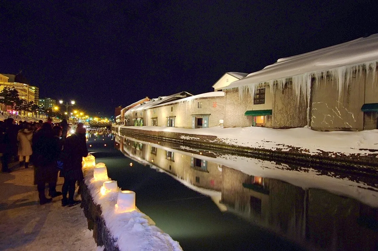A row of glowing snow lanterns along a snowy canal railing, with visitors walking by.