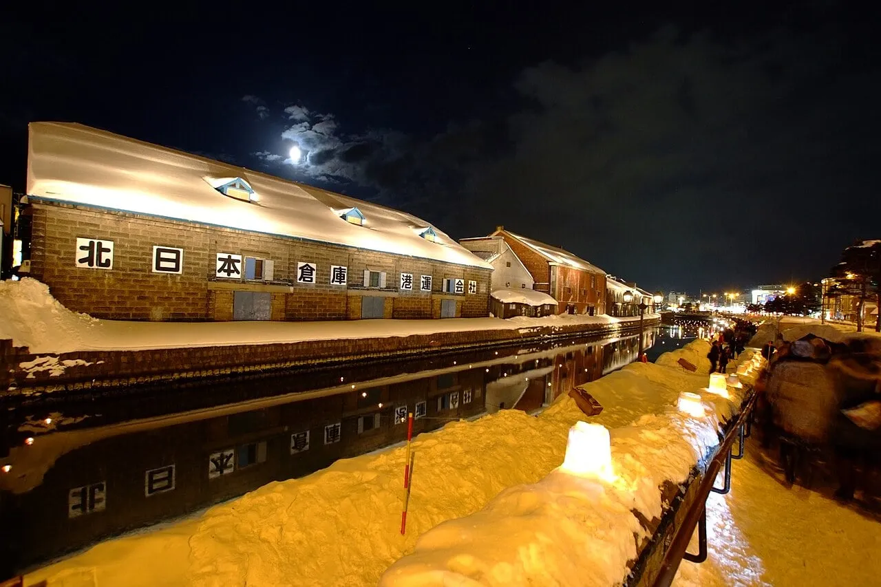 Snow lanterns line the canal path in front of lit stone warehouses under a moonlit sky.