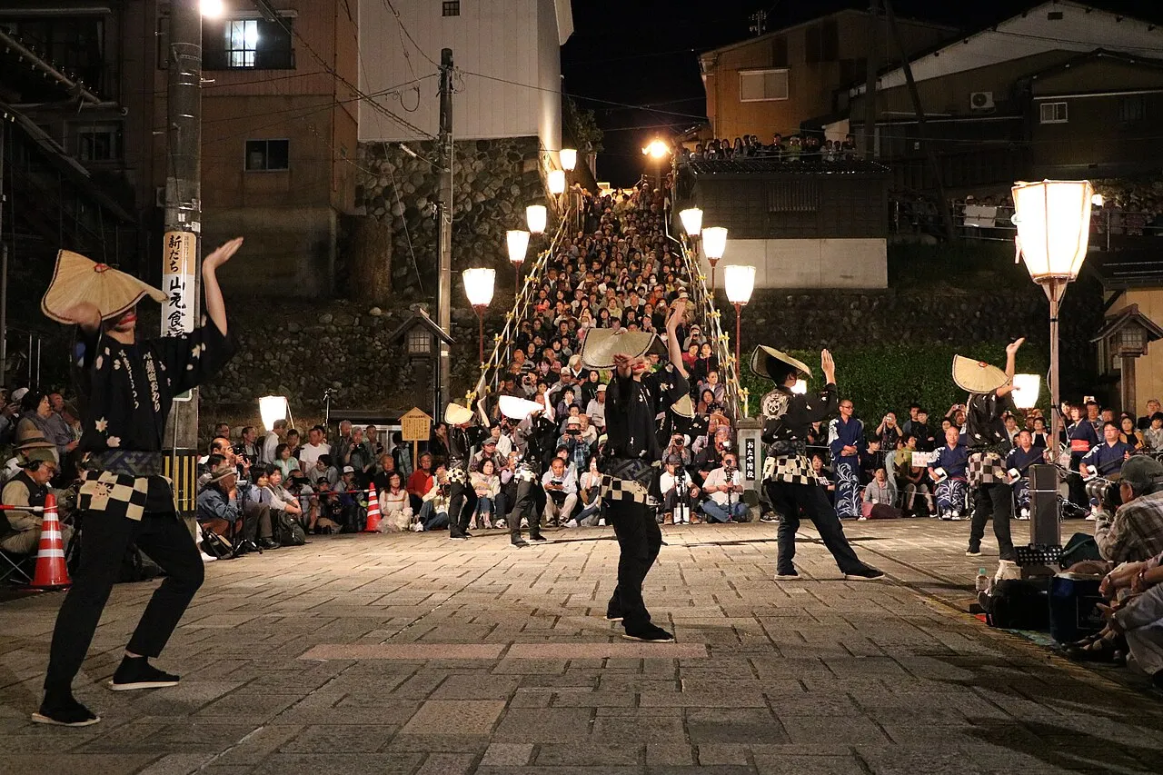 Dancers in straw hats perform on a lantern-lit stone square at night, with spectators filling a long stairway behind.