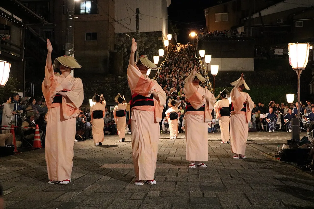 Women in pale kimono and deep straw hats dance in a line under street lanterns.