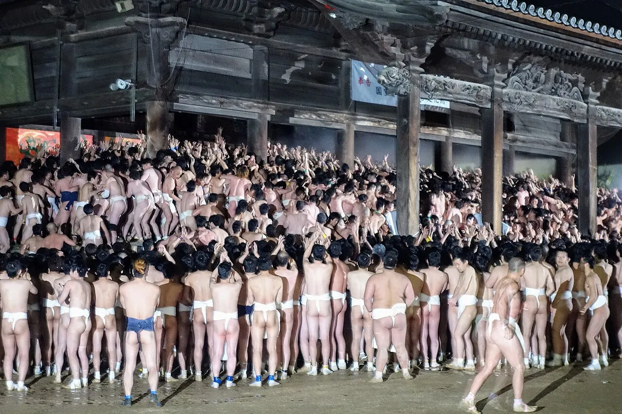 Hundreds of men in white loincloths packed tightly in front of a temple hall at night.
