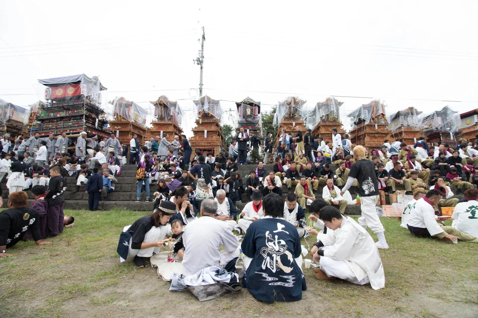 Danjiri floats lined up on a grassy riverbank above crowds of carriers in festival dress.