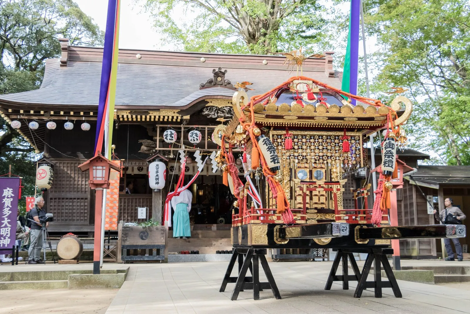 An ornate mikoshi stands on stands in front of the wooden hall of Makata Shrine.