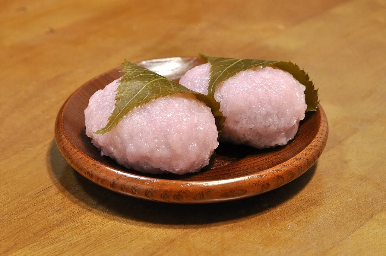 Two pink rice cakes wrapped in pickled cherry leaves on a wooden dish.