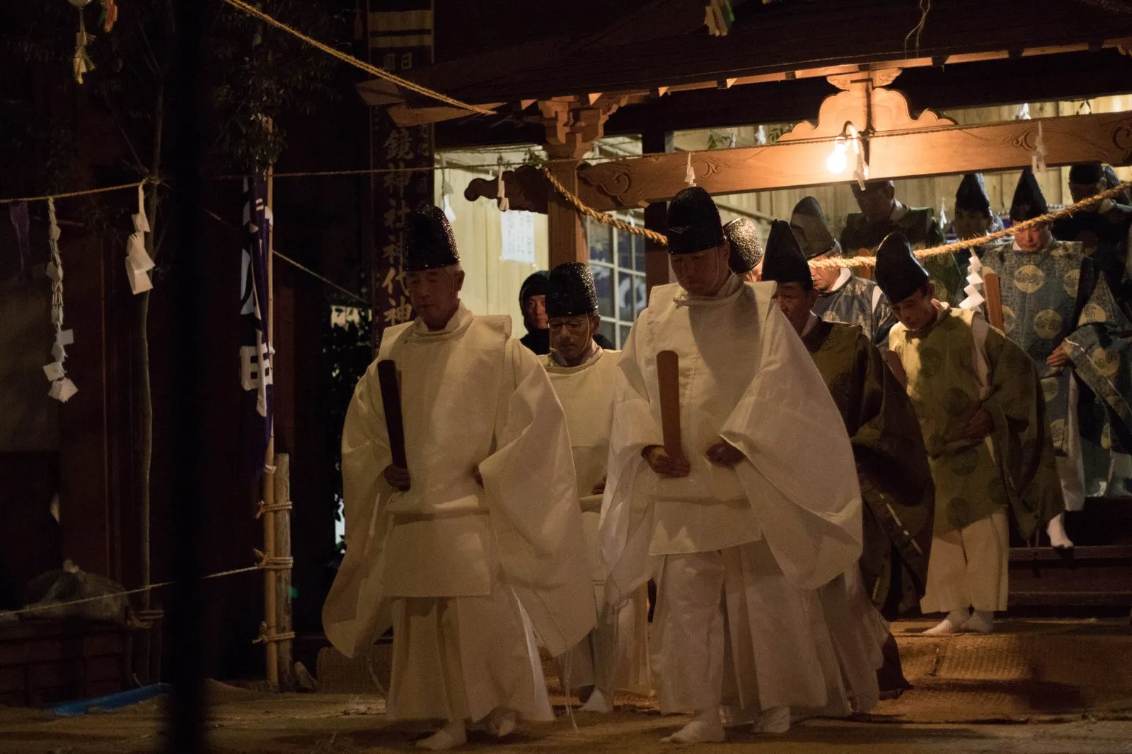 Performers in white robes and tall black hats dance inside a dim wooden shrine hall.