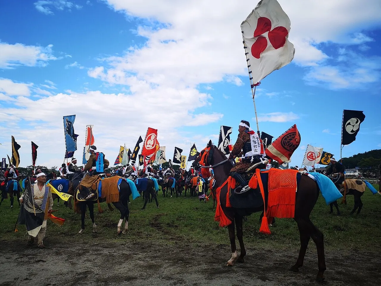 Riders in samurai armour with tall coloured banners on their backs sit on horses in a wide field under a blue sky.