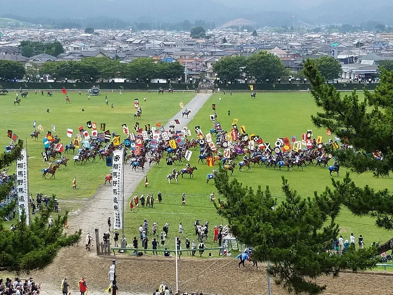 Seen from a hill, hundreds of riders with banners crowd a large green field ringed by spectators.