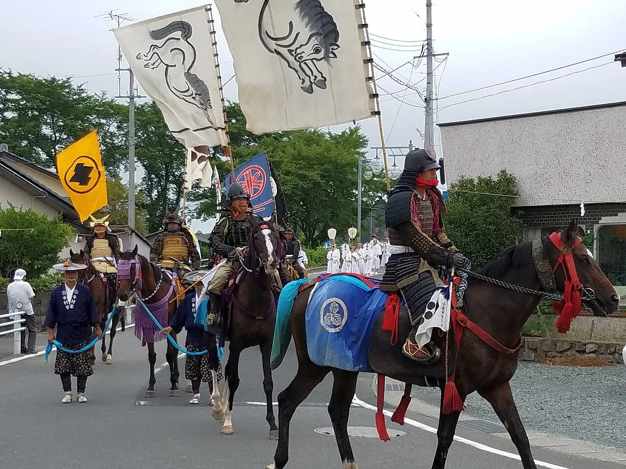 Mounted samurai in armour ride down a street with tall white banners on their backs.
