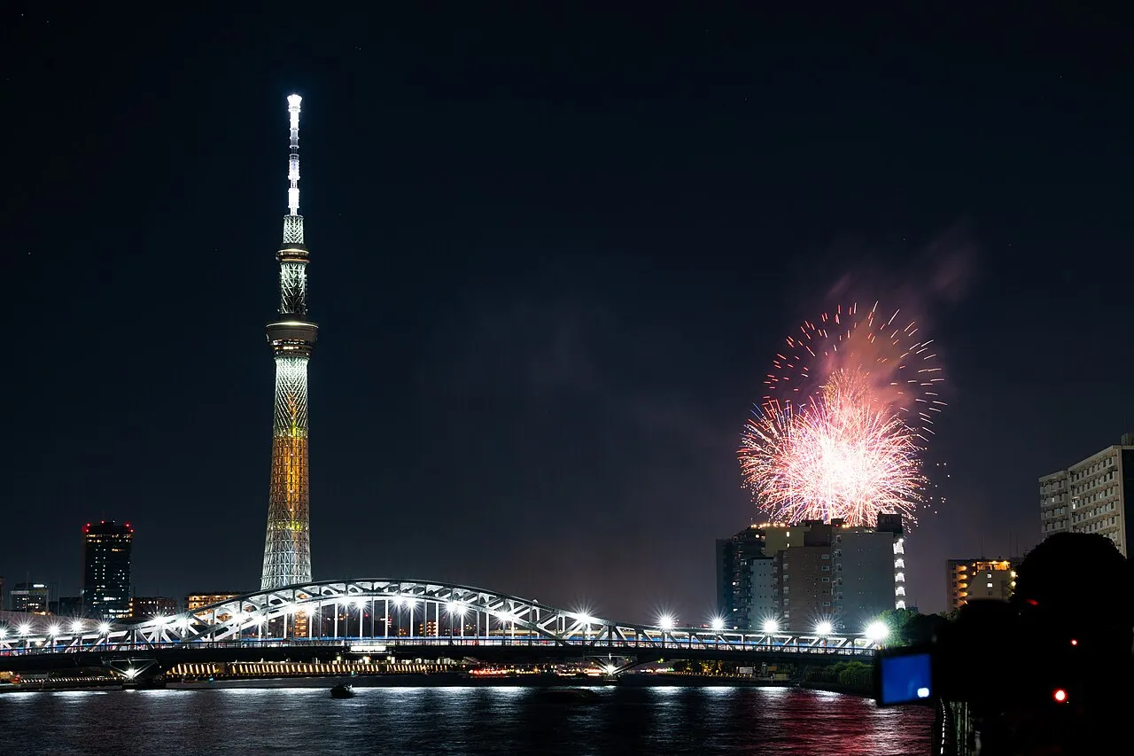 Fireworks burst beside the lit Tokyo Skytree, above an illuminated bridge over the Sumida River.