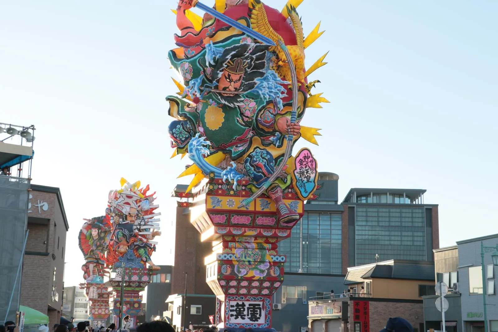 A painted warrior figure towers above the buildings on top of a tall float, with a second float behind it.