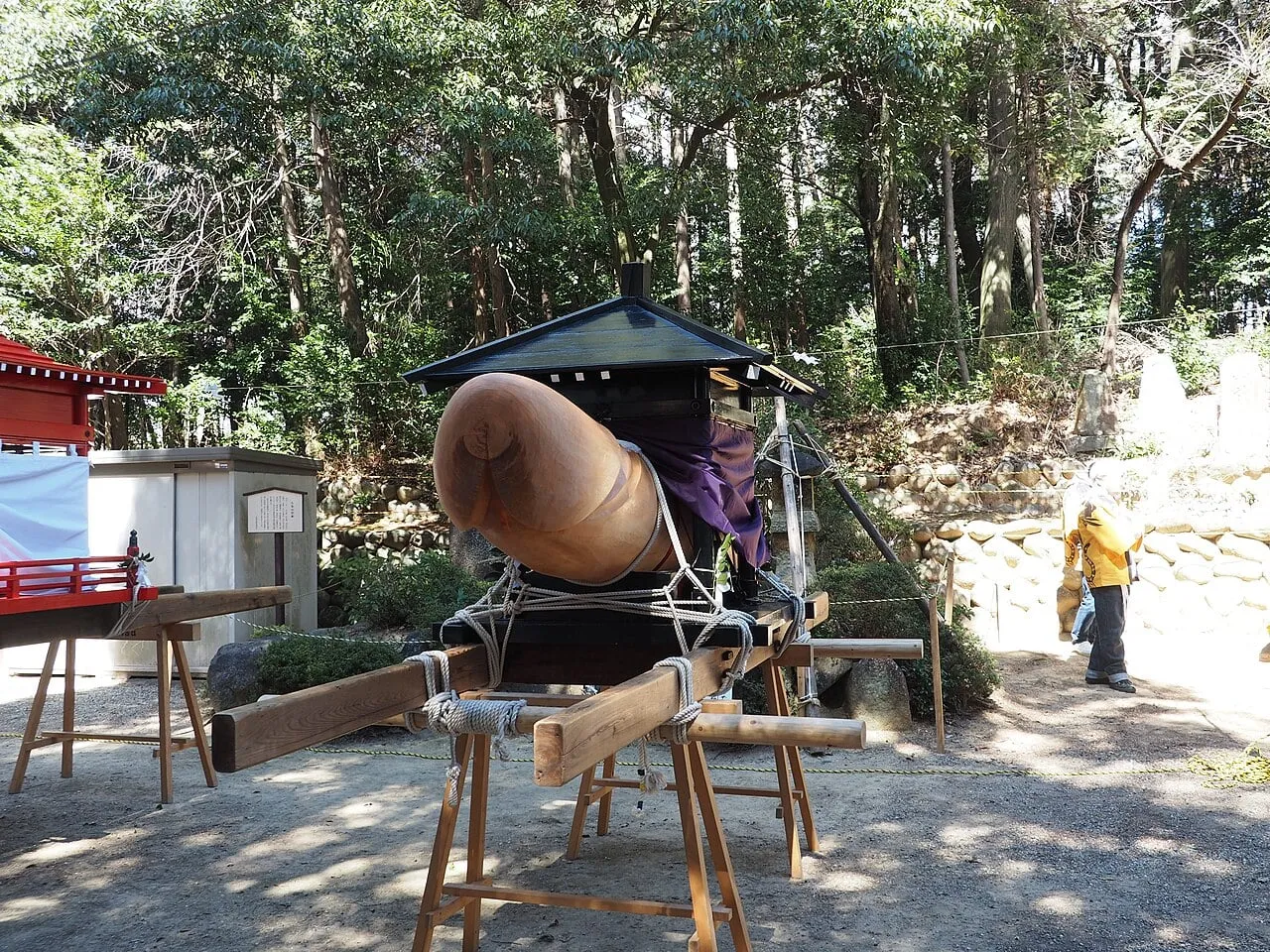 A huge wooden phallus on a black mikoshi under a small roof, resting on trestles among trees.