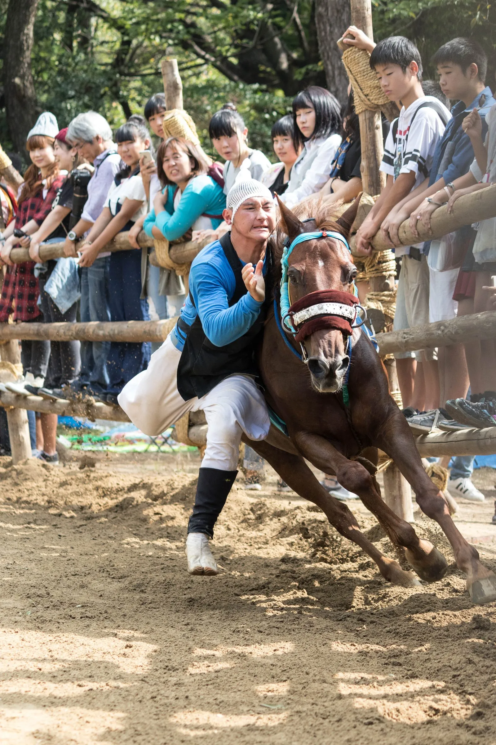 A young man in a blue happi coat runs alongside a galloping horse inside a fenced ring as spectators watch.