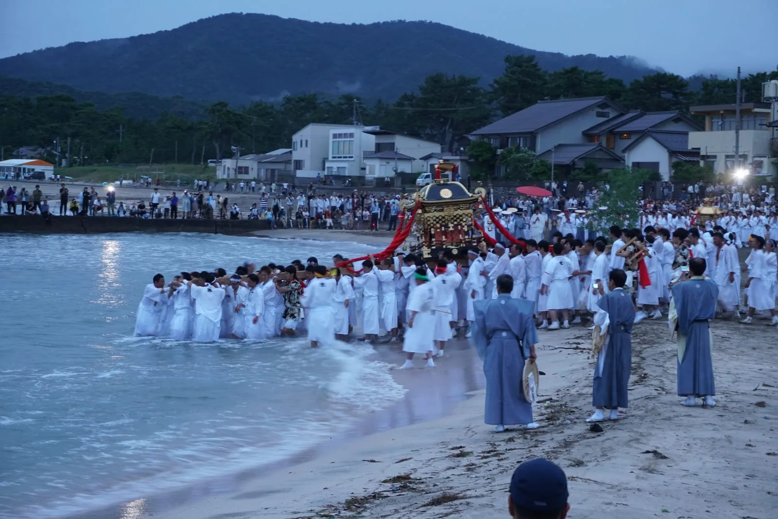 Men in white carry a portable shrine into the sea at dusk, watched by crowds on the beach.
