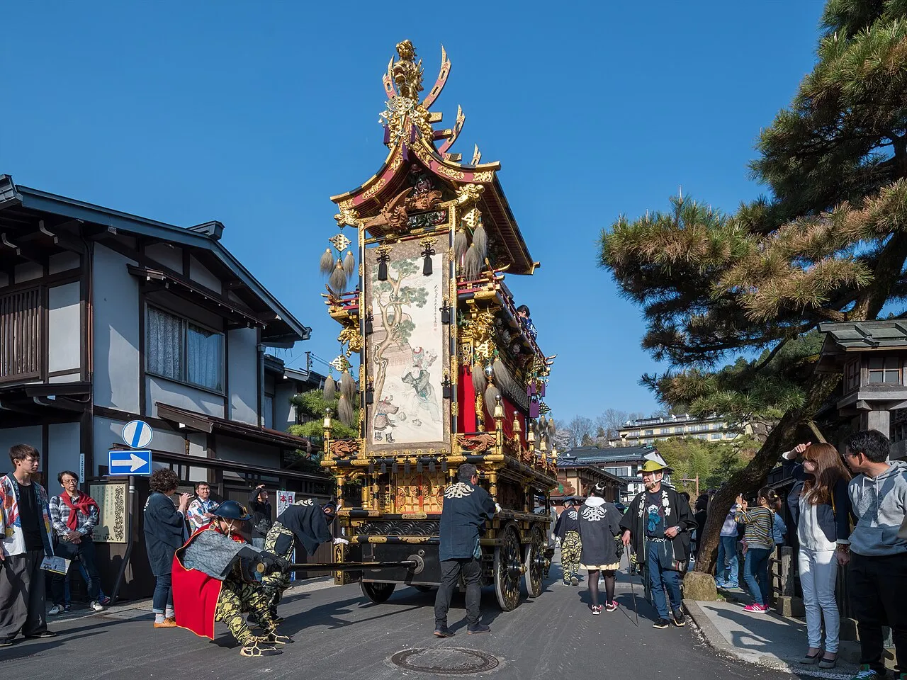 An ornate Takayama festival float with a carved gold roof stands in a street of old wooden houses, surrounded by people.