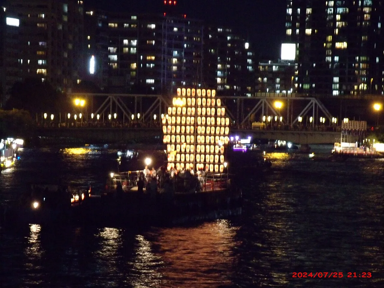A boat hung with glowing lanterns on the dark Ōkawa River, with city lights and a bridge behind.