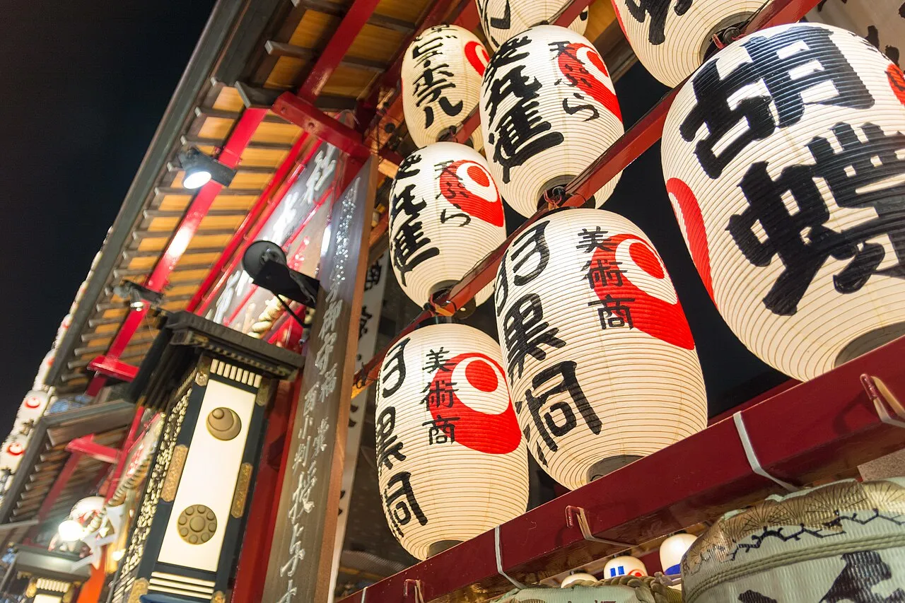 Large paper lanterns with black and red lettering hang above a stall at a shrine at night.