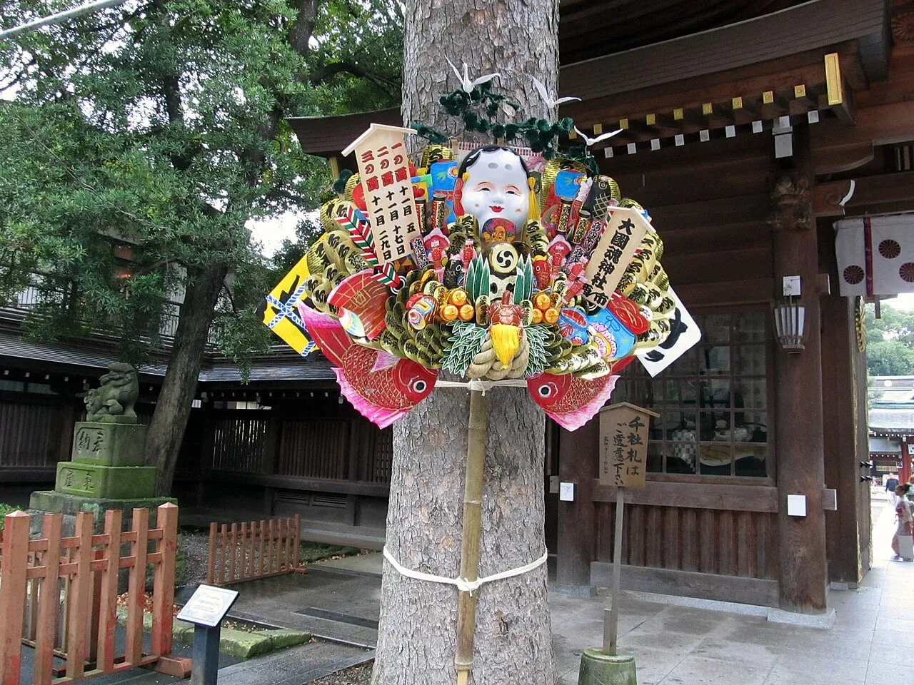 A large decorated bamboo rake covered with a mask of a smiling woman, gold coins, a sea bream and other lucky charms, tied to a tree.