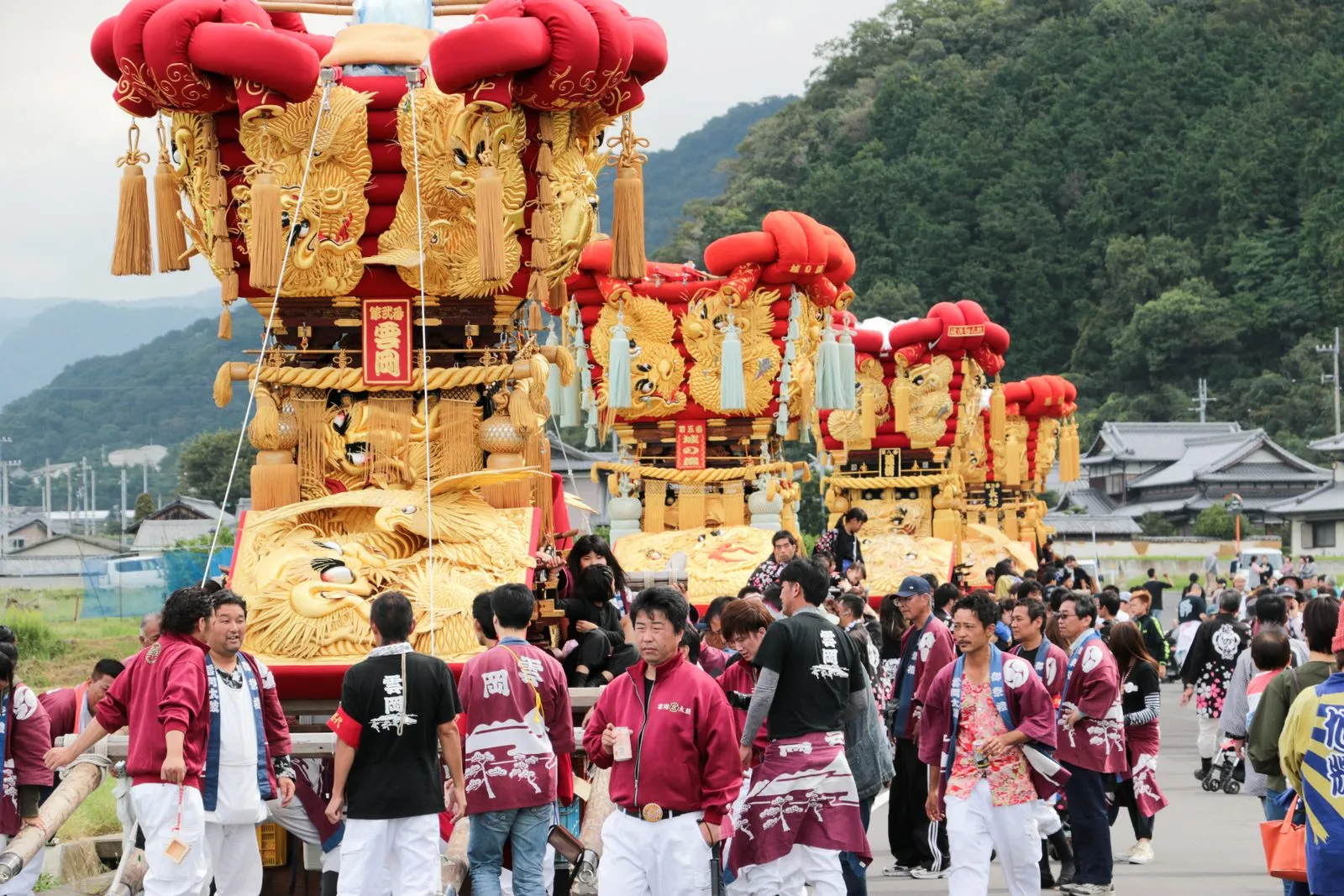 Gold-embroidered drum floats with red cushions carried by teams in maroon coats, with green hills behind.
