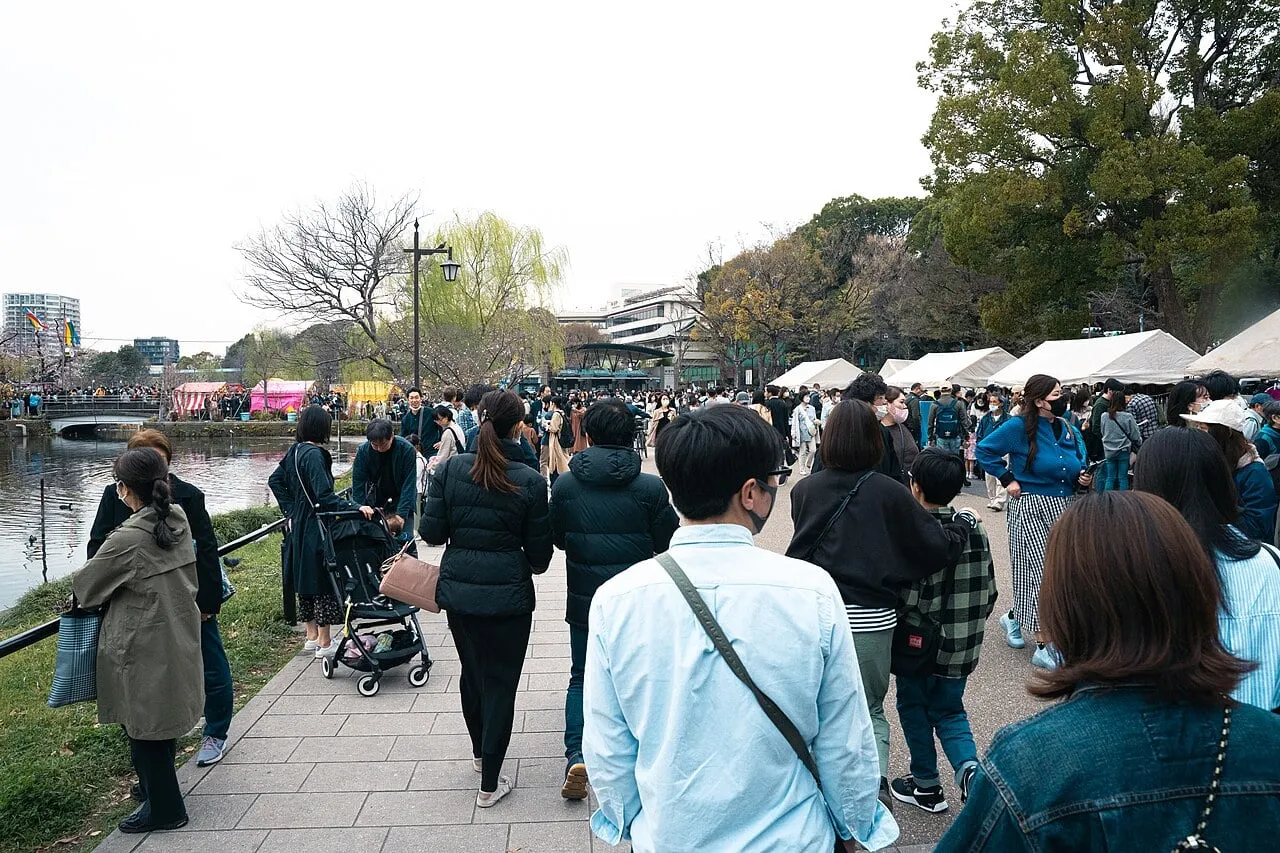 Crowds walk along a park path lined with food stalls and trees beside a pond.