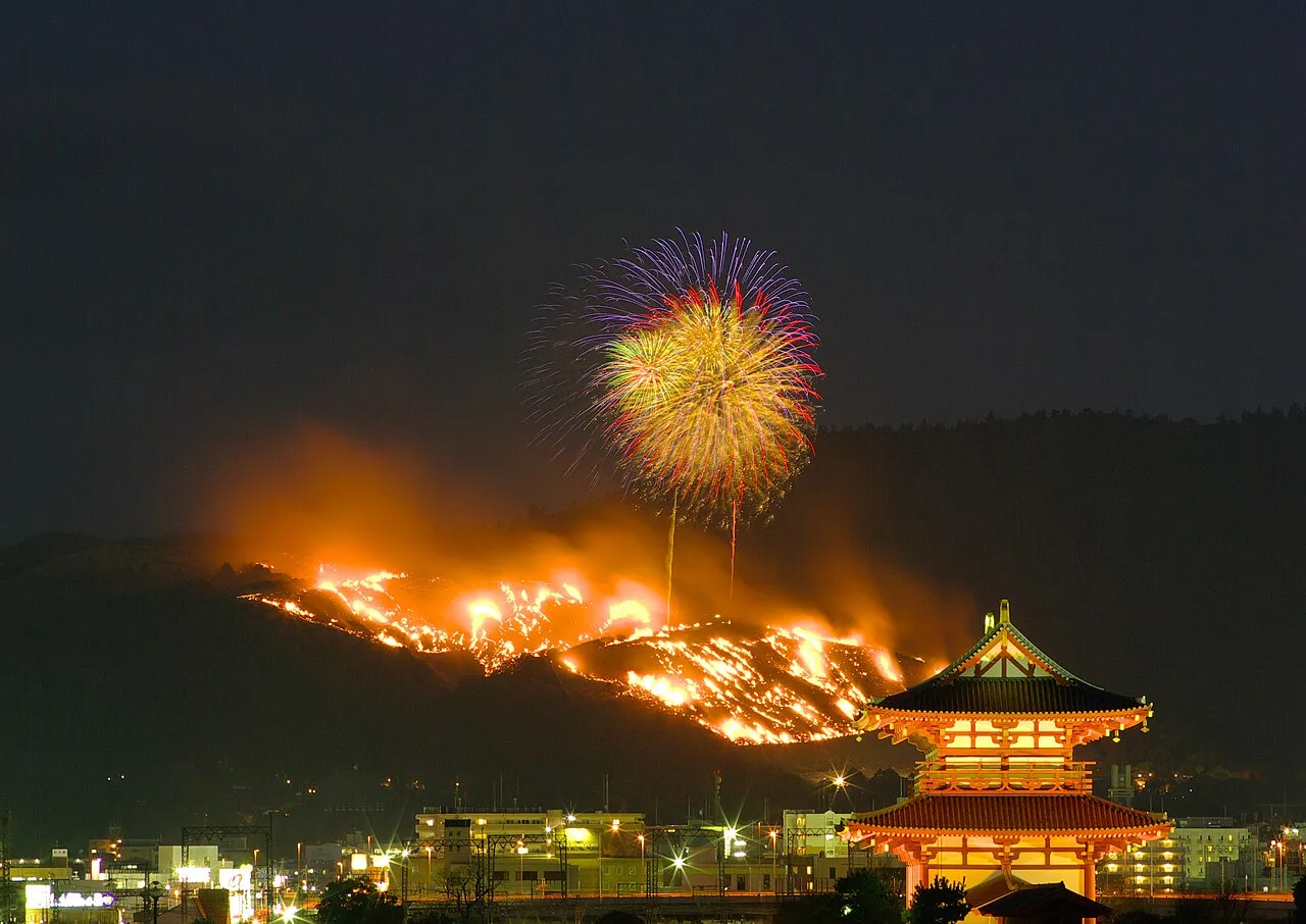 Fireworks burst over Mount Wakakusa as flames spread across the hillside, with the lit Suzakumon gate of the Heijō Palace site in front.