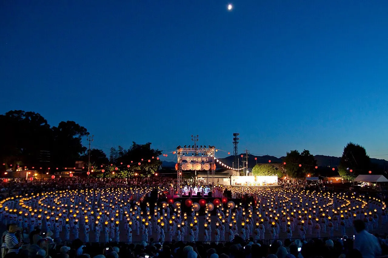 Hundreds of glowing lanterns arranged in circles on a field at dusk, with a lit stage in the center.
