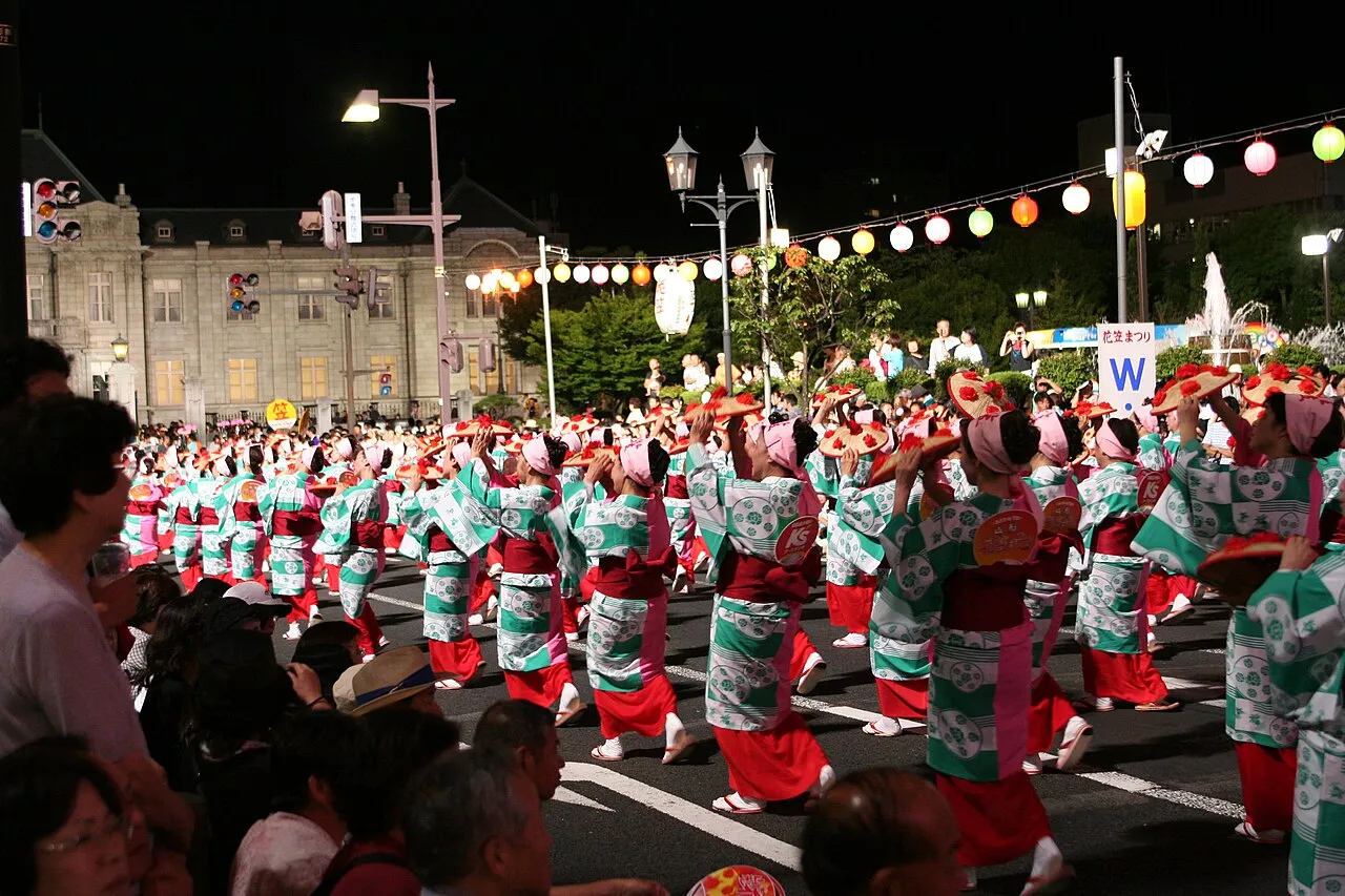 Rows of dancers in teal and red yukata twirl flower-decorated straw hats on a lantern-lit street at night.