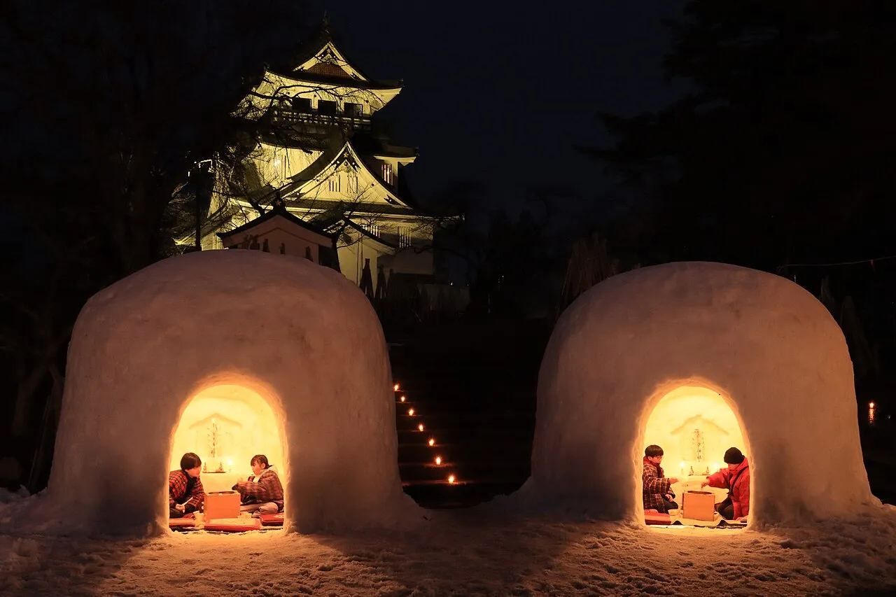 Two snow huts glowing from inside at night, with children seated within and Yokote Castle lit up behind.