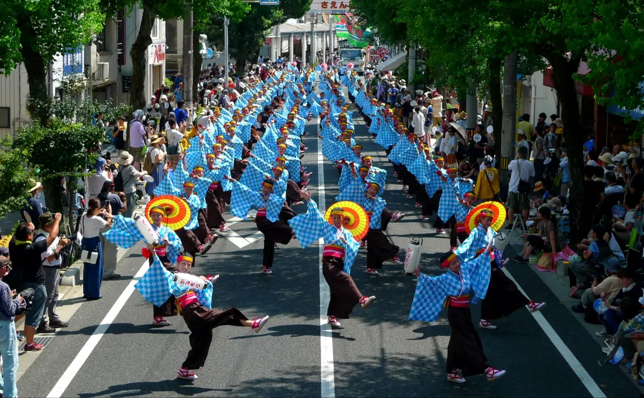 Rows of dancers in matching blue costumes with red-and-yellow parasols dance down a tree-lined street, seen from above.