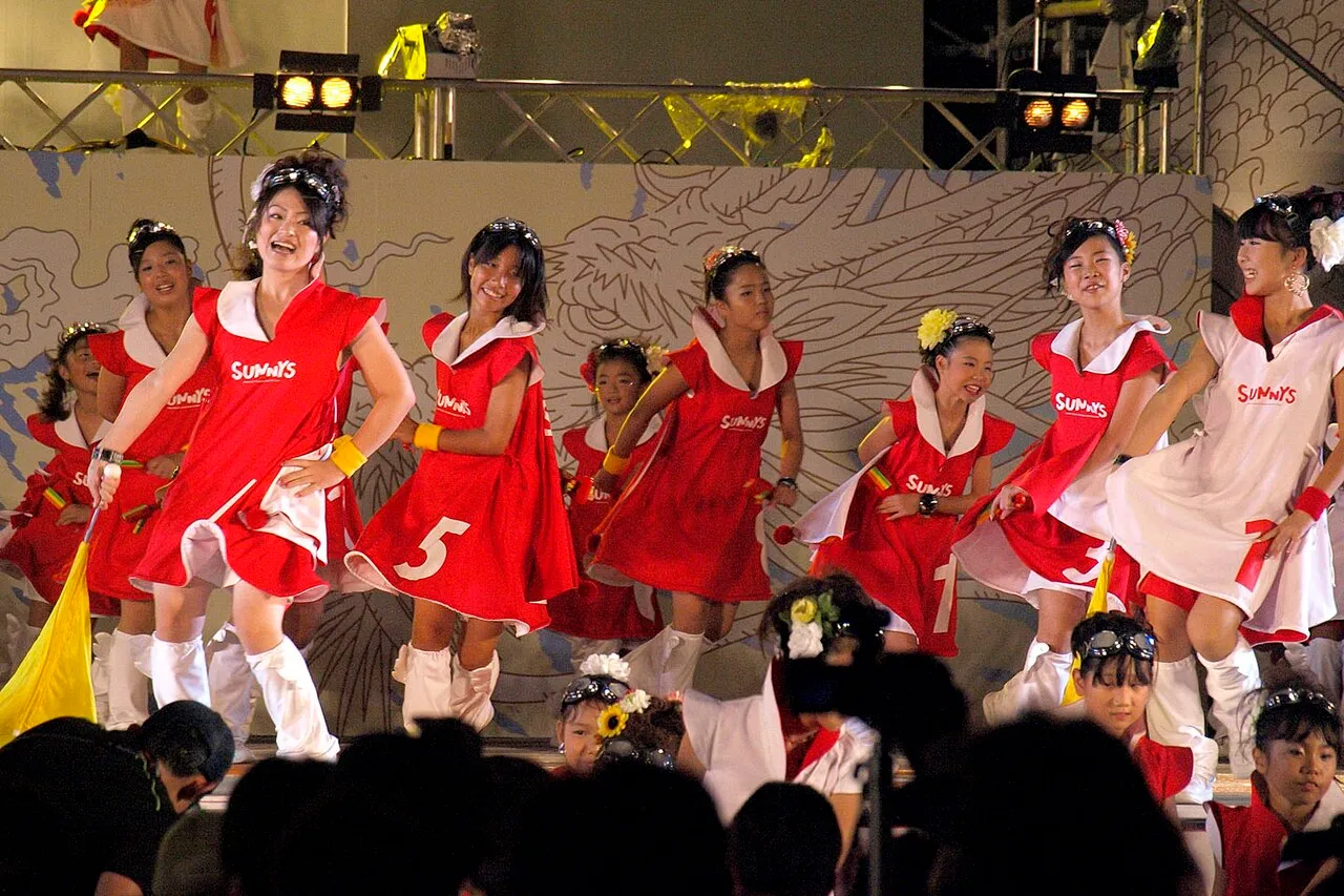 A team of dancers in red costumes with clappers performs on an outdoor stage in front of a crowd.