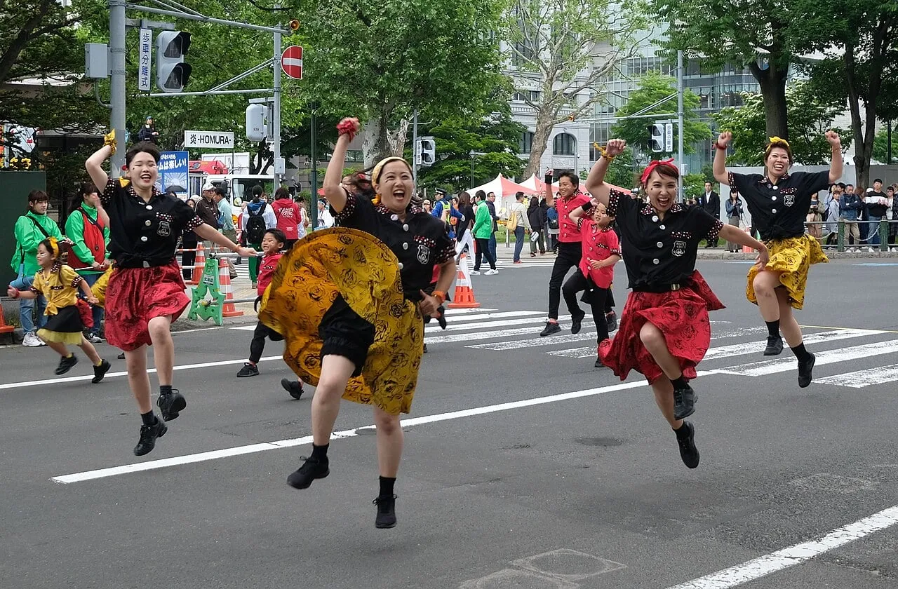 Dancers in black and red costumes leap down a city street with clappers in their hands.