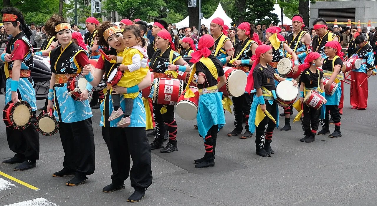 A large team in colourful costumes with drums and flags performs in a street lined with trees.