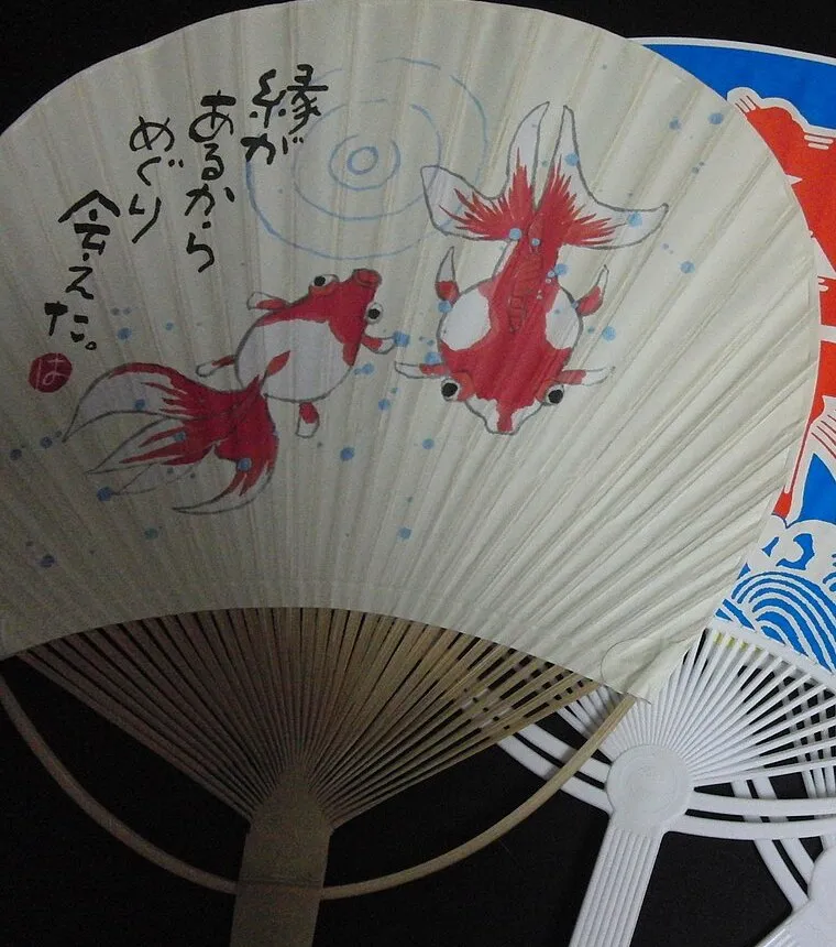 A round paper fan on a bamboo frame, painted with two red goldfish and a line of Japanese text.